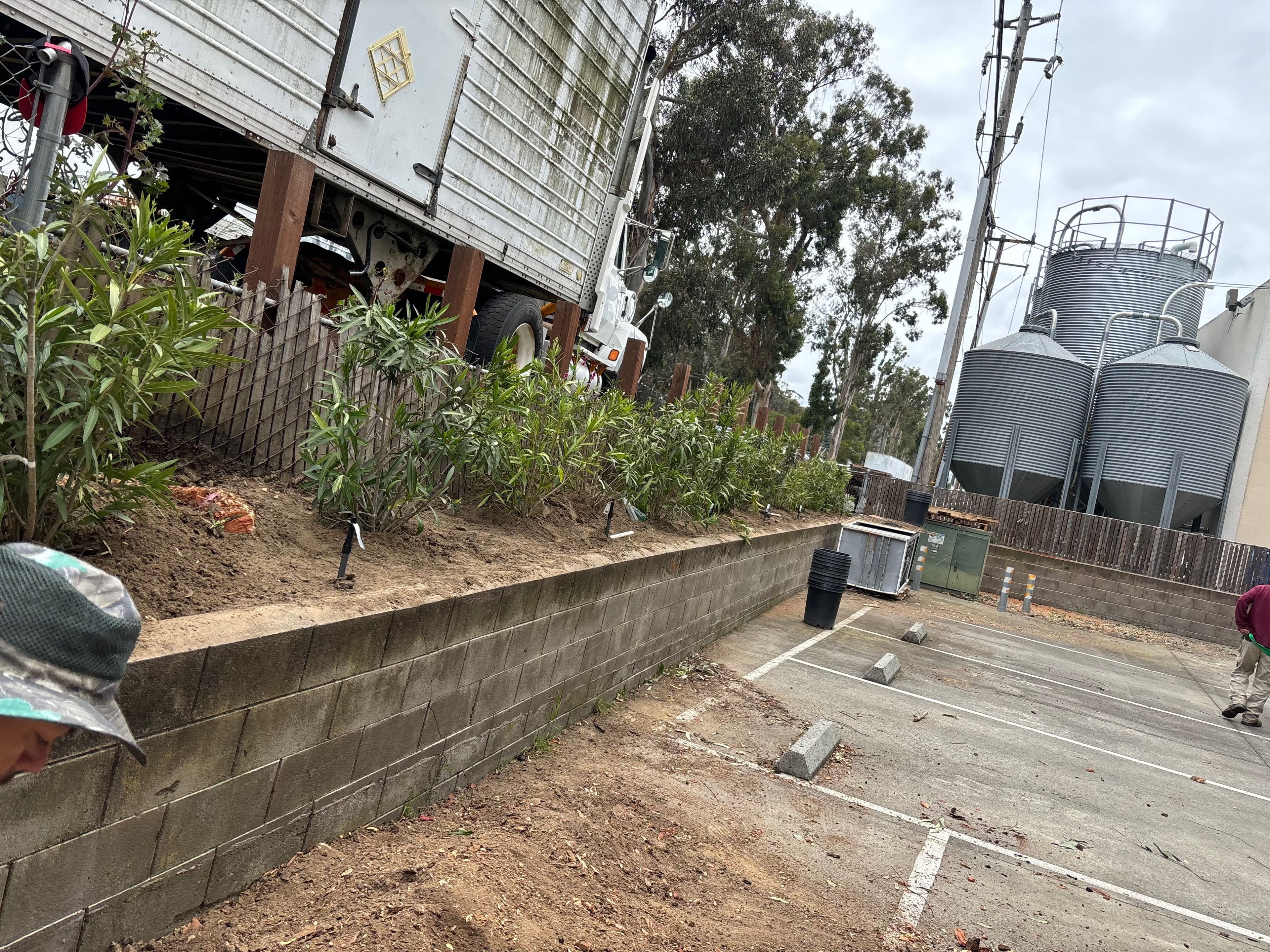 Parking lot with a wall, plants, and industrial silos in the background on a cloudy day.