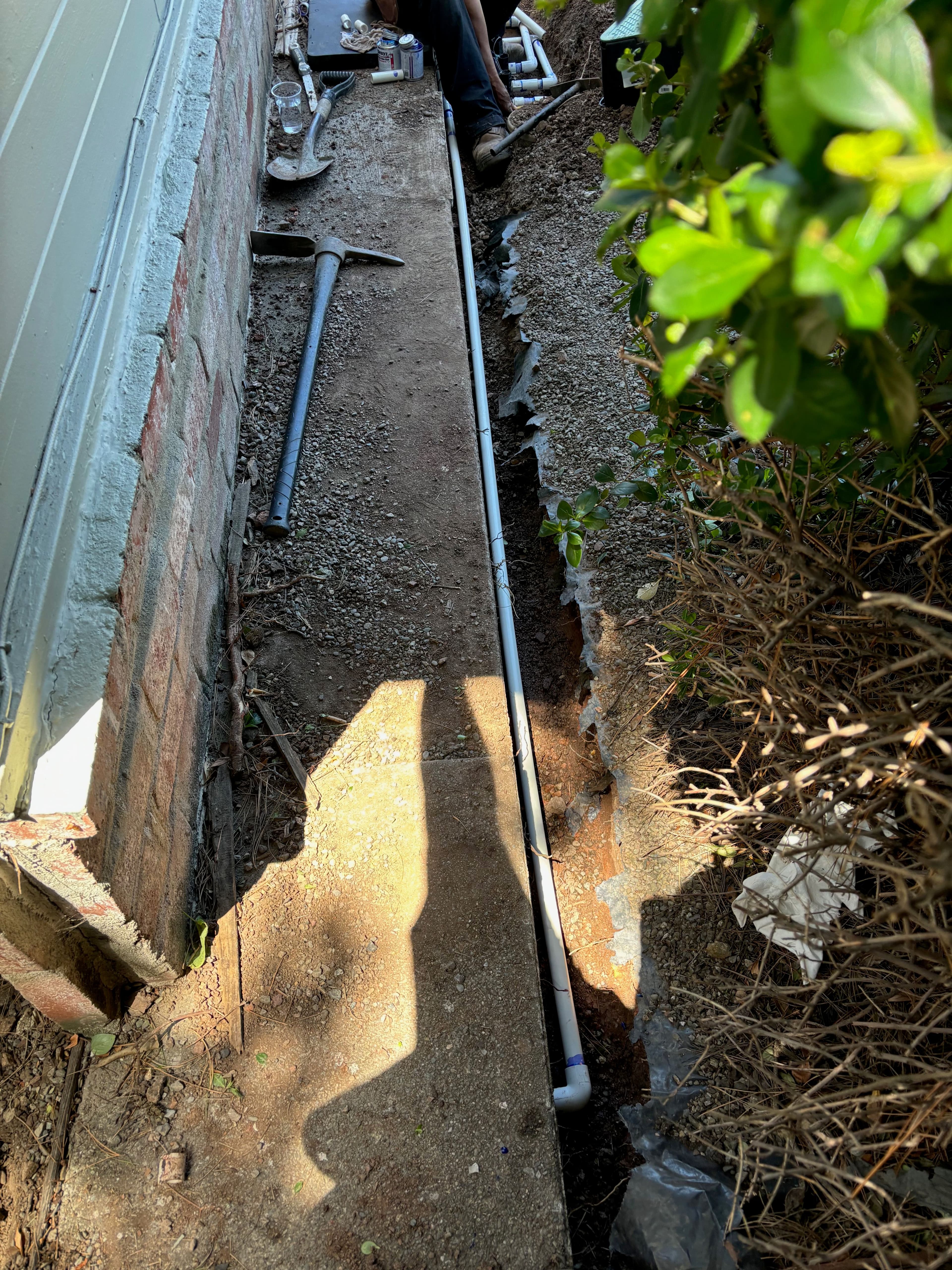 Person installing plumbing pipes in a trench beside a building with tools nearby.
